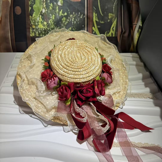 A beautifully detailed Evelina Victorian Romance straw boater hat, showcasing its coiled straw crown, a garland of red and pink roses with pearls, and a cascade of burgundy and lace ribbons, presented on a white surface.