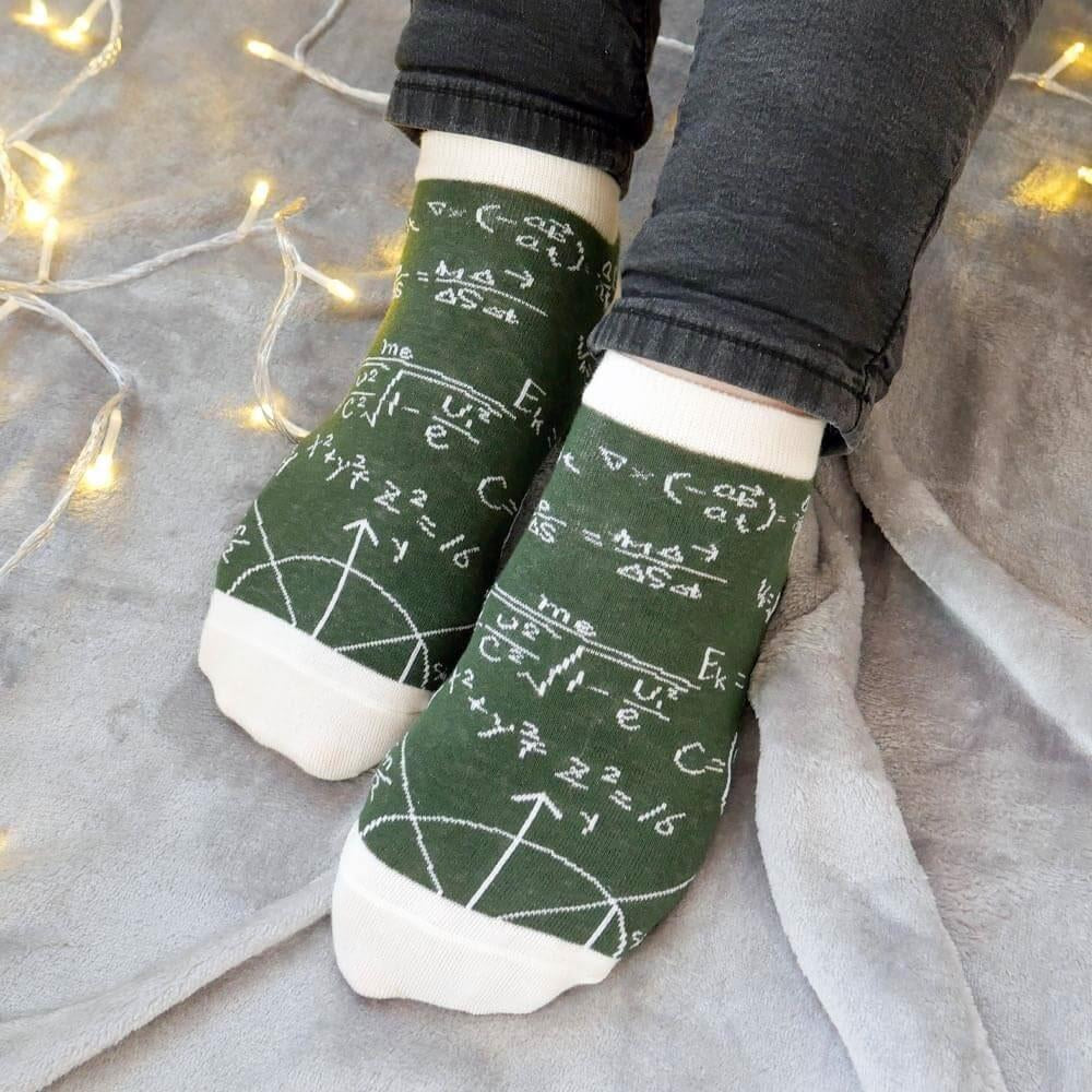 Green socks with mathematical equations worn by a person on a light gray surface with string lights.