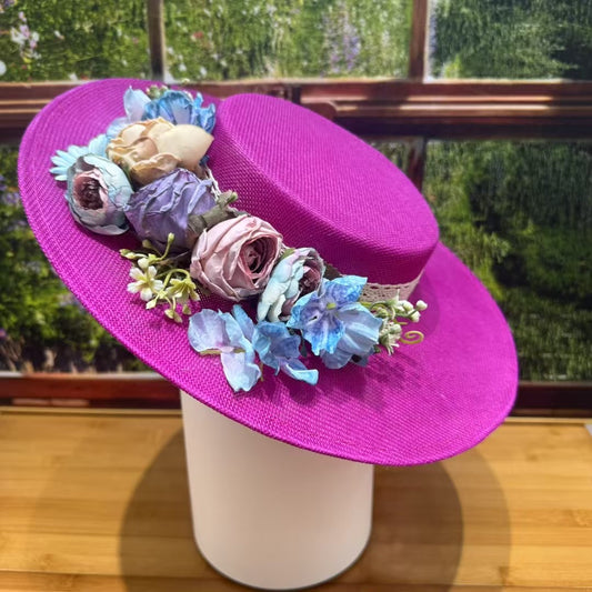 A vibrant fuchsia pink linen boater hat with a flat top, angled on a white stand. The crown is decorated with a ring of silk flowers in shades of blue, cream, and dusty rose, with a hint of white lace underneath.