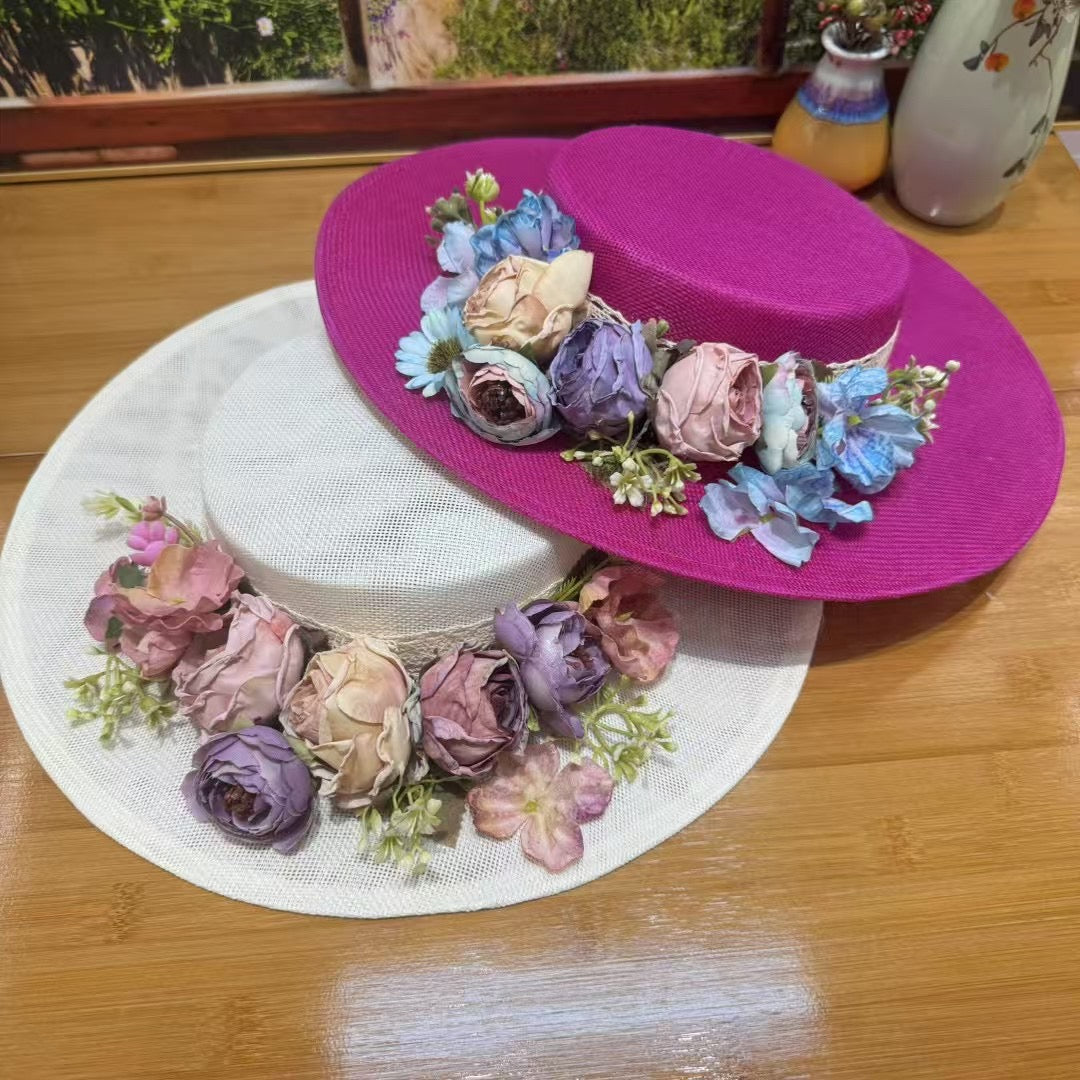 A vibrant fuchsia pink linen boater hat with a flat top, angled on a white stand. The crown is decorated with a ring of silk flowers in shades of blue, cream, and dusty rose, with a hint of white lace underneath.