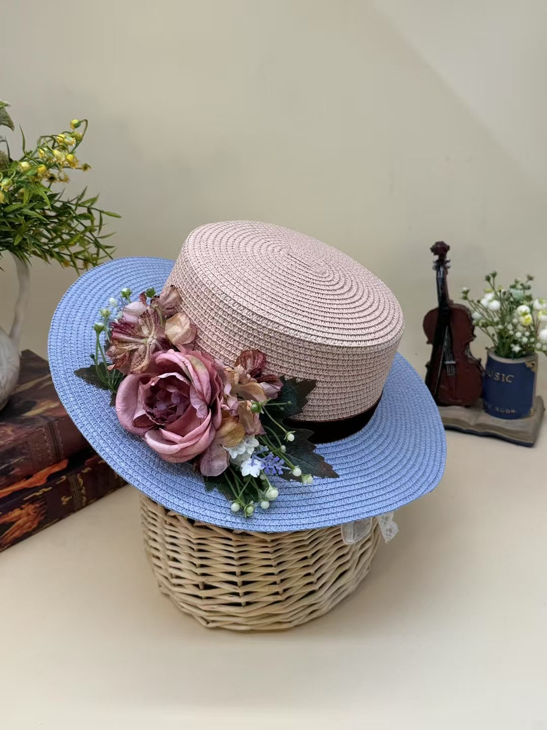 Vintage pink and blue floral boater hat made of papyrus with mauve roses and greenery, displayed on wicker basket with rustic decor.