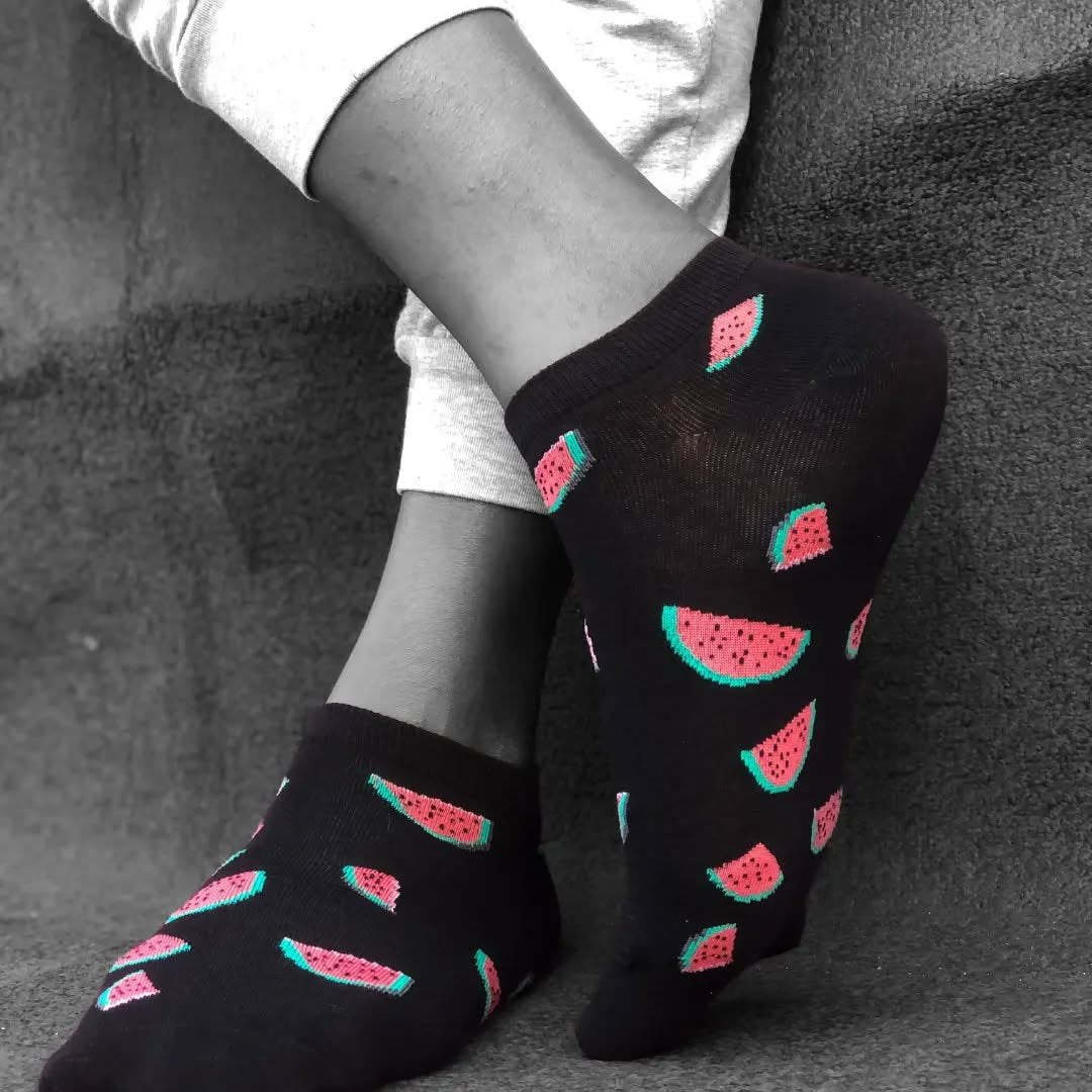 Black socks with watermelon pattern worn by a person sitting on a textured surface.