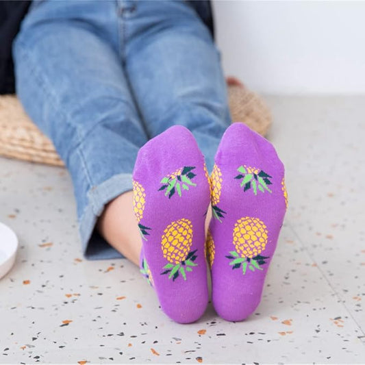 Purple socks with a quirky pineapple design worn by a person sitting on a speckled surface.