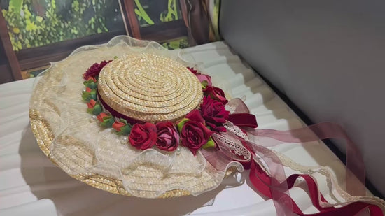A detailed video showcasing a Victorian-style straw boater hat. The camera pans around the hat, highlighting the coiled straw texture, the red and pink fabric roses, the lace-trimmed brim, and the long, flowing ribbons. A hand wearing a white lace glove gently handles the hat, turning it over to reveal a metal hair clip attached to the inside for secure wear.