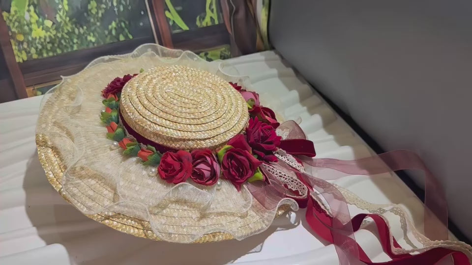 A detailed video showcasing a Victorian-style straw boater hat. The camera pans around the hat, highlighting the coiled straw texture, the red and pink fabric roses, the lace-trimmed brim, and the long, flowing ribbons. A hand wearing a white lace glove gently handles the hat, turning it over to reveal a metal hair clip attached to the inside for secure wear.